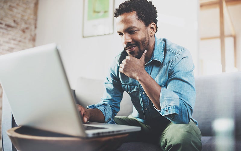 A man, sitting on his couch and engaging with a computer that's on a coffee table